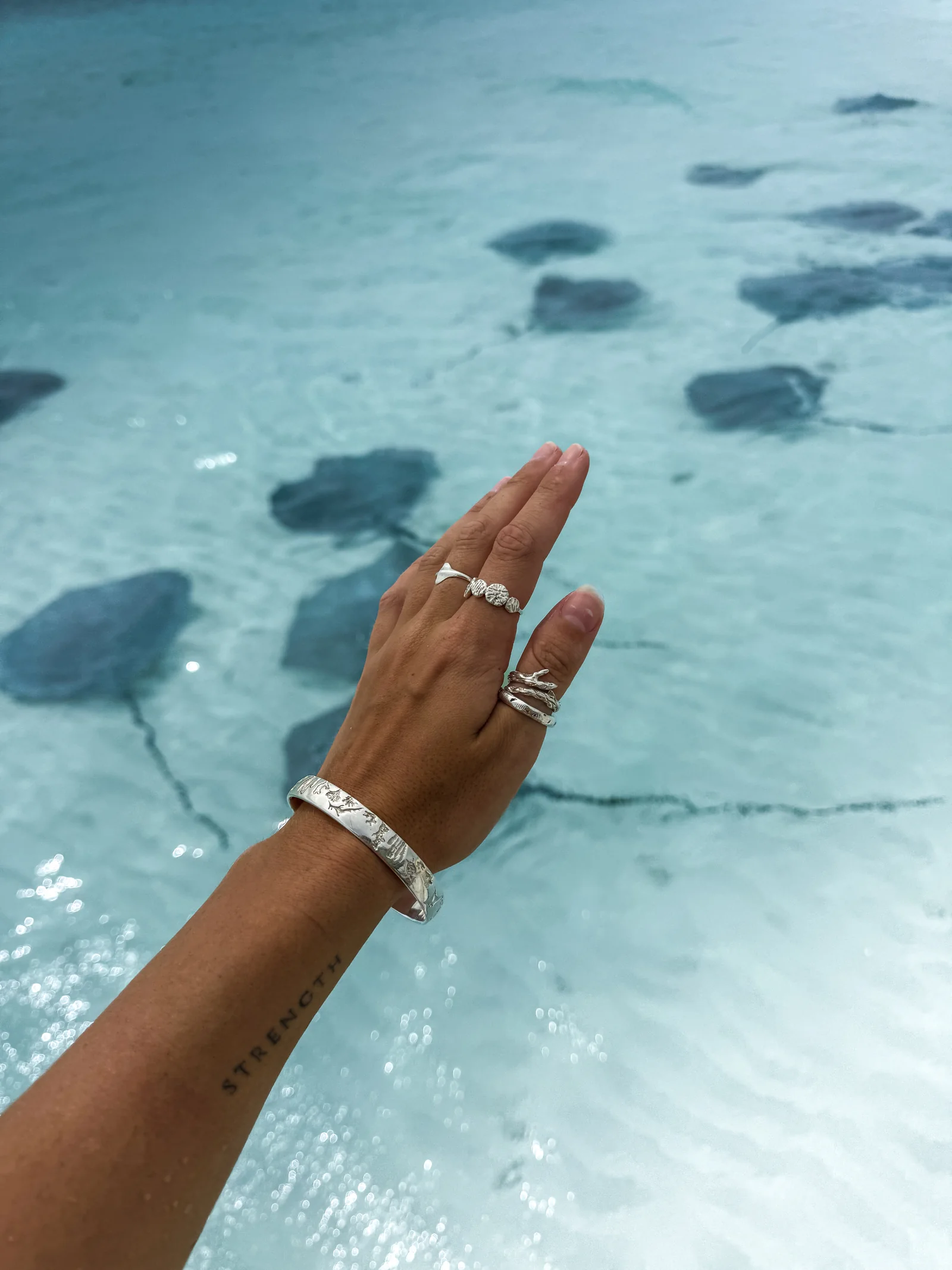 Hand wearing silver rings and a bangle above clear ocean water